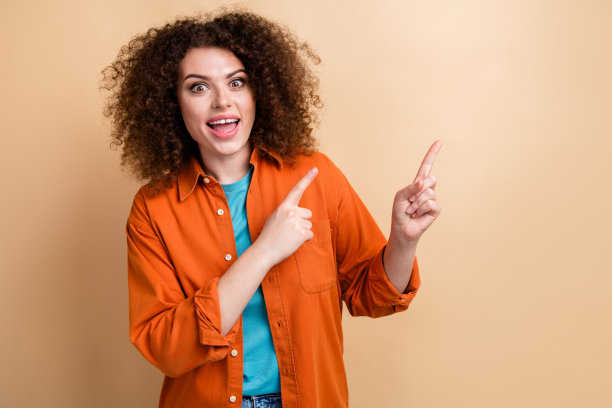 漂亮年轻女孩的照片肖像，兴奋地指向空白处，穿着时尚的橙色服装，背景为米色。图片下载