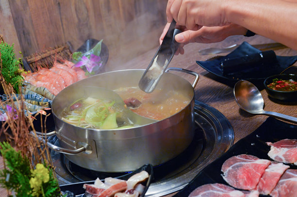 一个人的手用夹子将食材放入冒着热气的火锅中，桌上摆放着各种新鲜的肉类、海鲜和蔬菜。图片下载