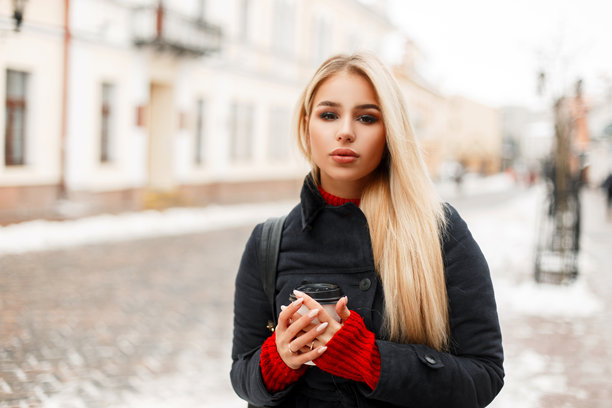 时尚年轻美女模特穿着复古风格的外套，手拿咖啡，在城市户外漫步。图片下载