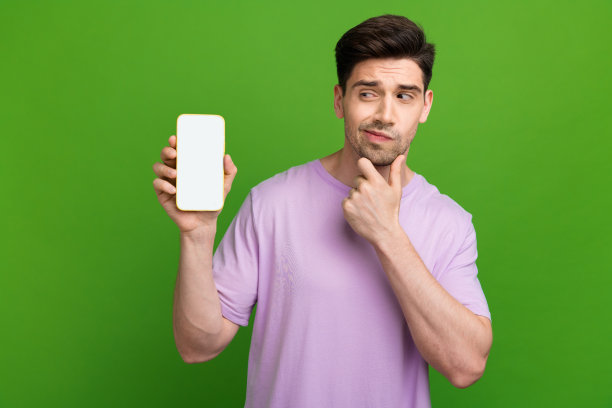 吸引人的年轻男士在绿色背景下，怀疑地用手触摸下巴，穿着时尚的紫色衣服，手持设备的照片肖像。图片下载