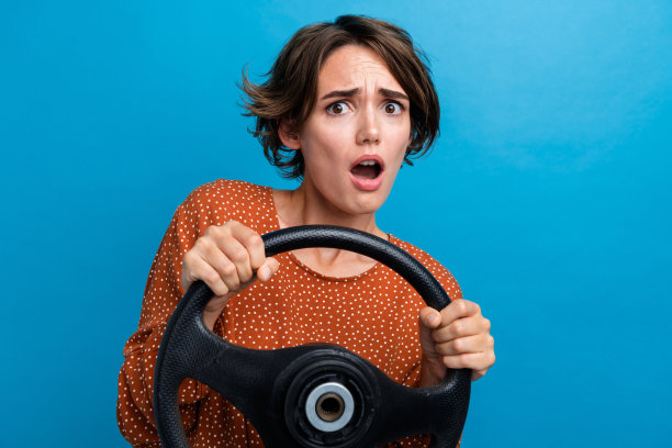 吸引人的年轻女性在蓝色背景下，穿着时尚的棕色衣服，面露恐惧反应，手握方向盘的照片肖像。图片下载