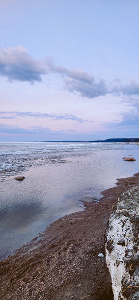 春季风景。美丽的黎明全景，海湾上方的积云在明亮的光线下。海岸线上有冰、雪和岩石。阳光的光芒在海面上反射。竖版照片图片下载