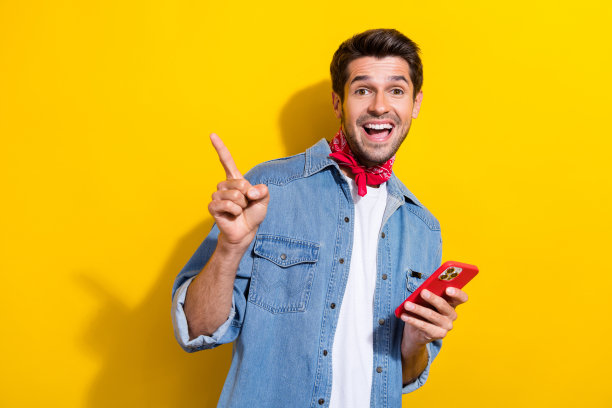 吸引人的年轻男子在黄色背景下穿着时尚牛仔裤，指向空白处，手持设备的照片肖像图片下载