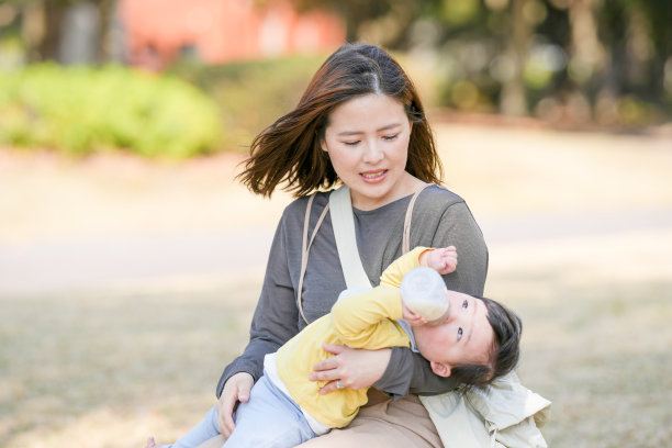 在日本的一个阳光明媚的森林公园里，一位三十多岁的日本妈妈坐在长椅上，正在给她六个月大的宝宝喂奶。这张照片捕捉了户外宁静而温馨的家庭时刻。图片下载