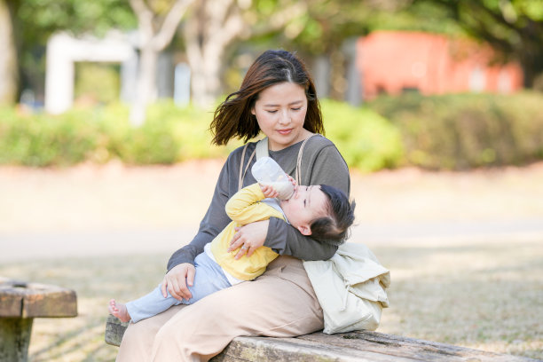 在日本的一个阳光明媚的森林公园里，一位三十多岁的日本妈妈坐在长椅上，正在给她六个月大的宝宝喂奶。这张照片捕捉了一个安静而温馨的户外家庭时刻。图片下载