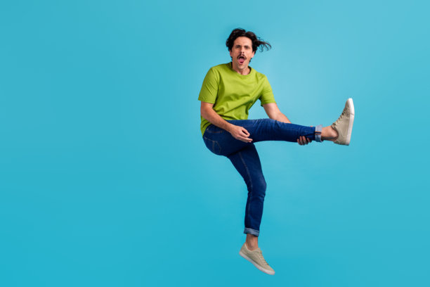 全身照片，帅气搞笑的男士穿着绿色T恤，跳得很高，玩着腿吉他，背景为蓝色。图片下载