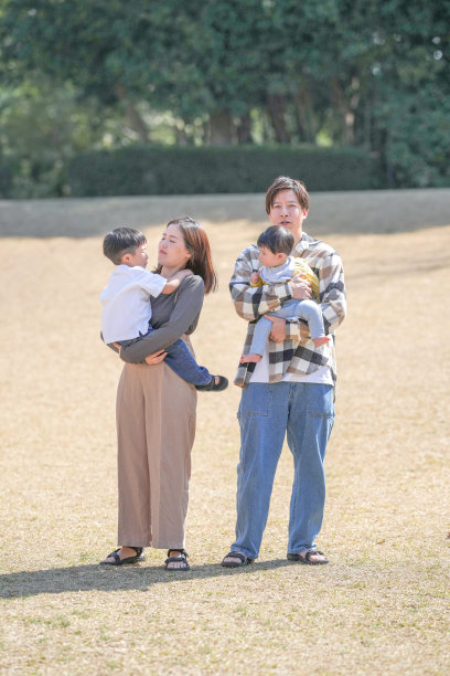 在日本的一个阳光明媚的森林公园里，一对日本夫妇与他们3岁的儿子和6个月大的宝宝站在一起。这张照片捕捉了一个宁静而温馨的家庭时刻，背景是一个宁静的公园环境。图片下载