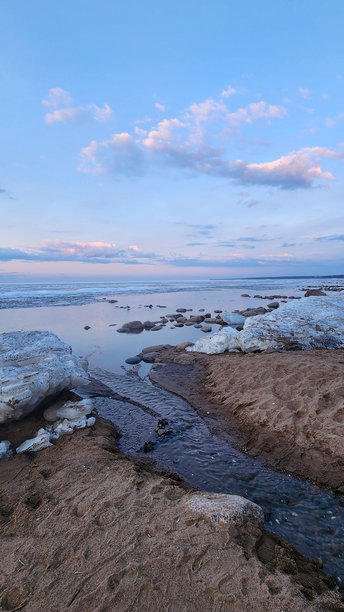 春季风景。美丽的黎明全景，海湾上方的积云在明亮的光线下。海岸线上有冰、雪和岩石。阳光的光芒在海面上反射。竖版照片图片下载