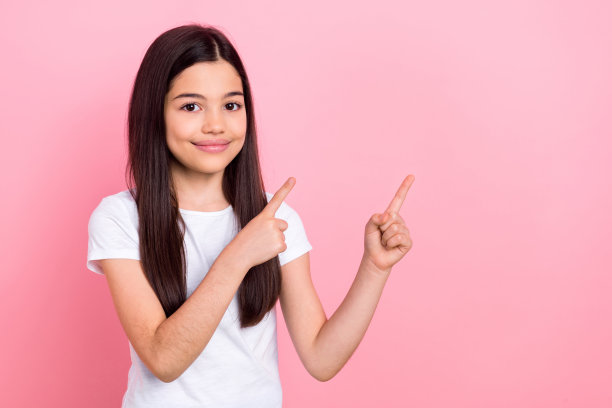 可爱的女学生在粉色背景上穿着时尚的白色衣服，手指指向空白处的照片肖像。图片下载