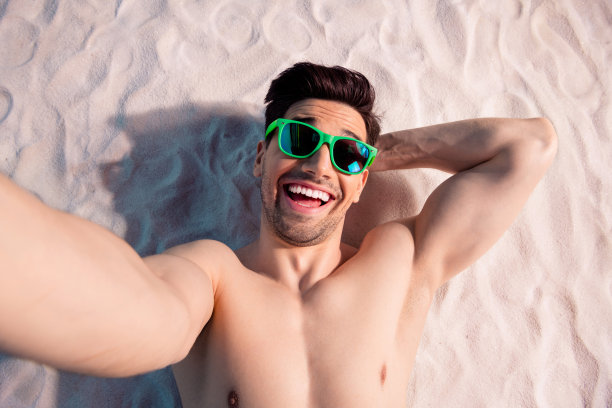 梦幻兴奋的赤裸上身男子在海边沙滩上仰卧，手放在头后自拍的俯视照片图片下载