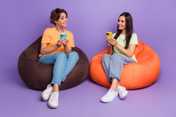 两位女孩坐在豆袋椅上，使用iPhone一起交流，背景为紫色，呈现全身长度的照片。图片下载