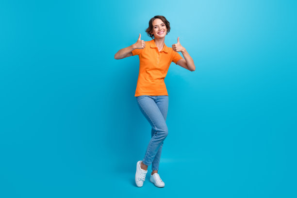 全身照片，积极开朗的女孩穿着时尚的马球衫和牛仔裤，竖起大拇指表示工作出色，背景为蓝色。图片下载