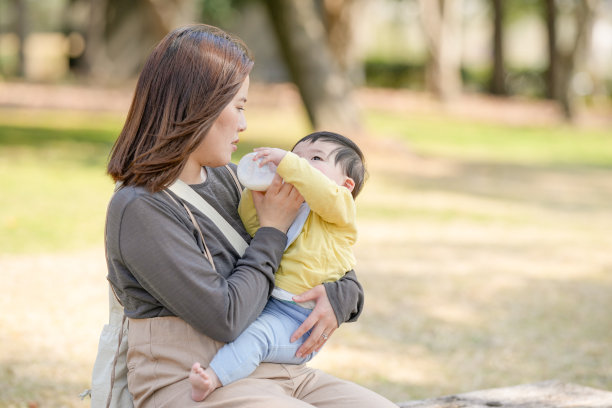 在日本的一个阳光明媚的森林公园里，一位三十多岁的日本妈妈坐在长椅上，给她六个月大的宝宝喂奶。这张照片捕捉了一个安静而温馨的户外家庭时刻。图片下载