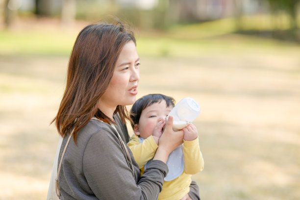 在日本的一个阳光明媚的森林公园里，一位三十多岁的日本妈妈坐在长椅上，正在给她六个月大的宝宝喂奶。这张照片捕捉了一个安静而温馨的户外家庭时刻。图片下载