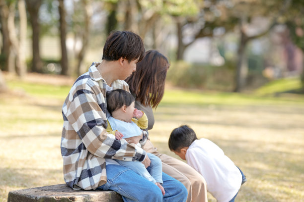 在福冈的阳光明媚的公园里，一对三十多岁的日本夫妇坐在长椅上，陪伴着他们三岁的儿子和六个月大的女儿。这张照片捕捉了一个宁静的家庭户外时刻。图片下载