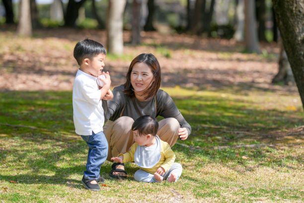 在日本的一个阳光明媚的森林公园里，一位日本妈妈和她六个月大的宝宝坐在草地上。附近，她三岁的儿子正在扔球。这张照片捕捉了一个宁静的春季家庭时刻。图片下载