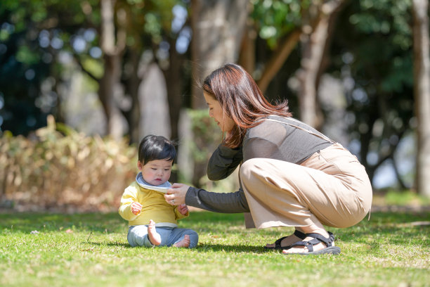 在日本的一个阳光明媚的森林公园里，一名6个月大的日本宝宝坐在草地上，旁边一位三十多岁的日本妈妈在注视着他。这张照片展现了一个宁静而充满爱的春日时刻。图片下载