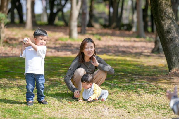 在日本的一个阳光明媚的森林公园里，一位日本妈妈和她六个月大的宝宝坐在草地上。附近，她三岁的儿子正在扔球。这张照片捕捉了一个宁静的春季家庭时刻。图片下载