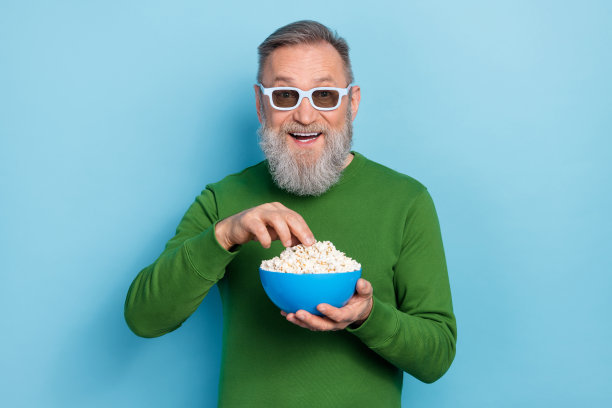 戴眼镜、穿毛衣的积极老年男子在蓝色背景下吃爆米花观看电影系列的照片图片下载