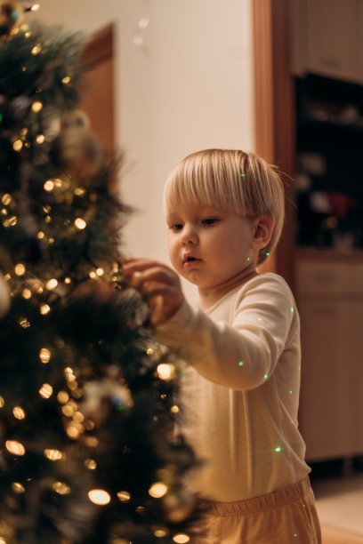 A toddler stands near a sparkling Christmas tree clapping joyfully as his smiling mother joins in from the bed capturing a warm festive moment filled with love celebration and connection图片下载