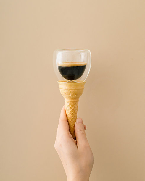 一张独特的概念照片，女性手握冰淇淋蛋筒，旁边有一杯咖啡，背景为米色。极简超现实的食品和饮料概念。时尚的冰淇淋创意。咖啡美学。图片下载