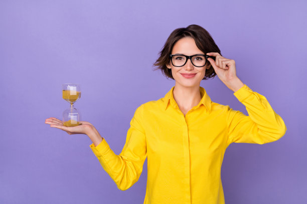 一位穿着黄色服装、佩戴眼镜的年轻女性在人紫色背景下手持时钟的照片图片下载