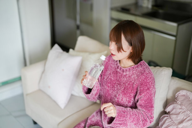 在一个整洁简约的房间里，一位二十多岁的日本孕妇，短棕色头发，穿着粉色针织裙，坐在沙发上，慢慢地从塑料瓶中喝水。图片下载