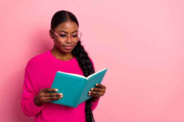可爱的千禧世代棕发女士穿着毛衣，戴着眼镜，坐在粉色背景下阅读书籍的照片图片下载