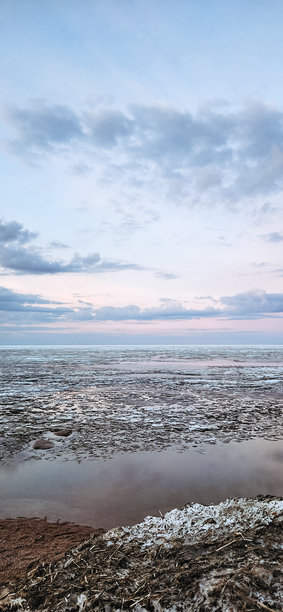 春季风景。美丽的海湾晨曦全景。明亮的光线下，水面上漂浮着积云。海岸线上有冰雪和岩石。阳光的光芒在海面上反射。竖版照片图片下载