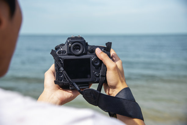 夏季海滩蓝天上，男旅行者使用数码相机拍照。亚洲摄影师在夏季的海滩上放松旅行。男背包客在亚洲目的地拍摄照片，享受假期之旅。图片下载