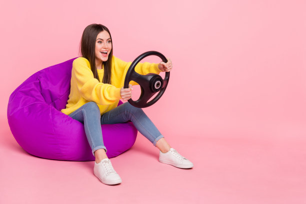 年轻女士坐在椅子上玩视频游戏，骑着车辆的全身侧面照片，背景为粉色。图片下载