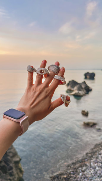 一只装饰着贝壳如同戒指的女人手在日落时分高举于平静的海面前。水中耸立着岩石，天空被柔和的晚霞渲染成粉彩色调。垂直旅行照片图片下载