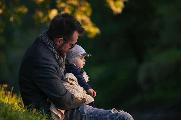 胡须父亲与婴儿儿子在公园草地上共度时光的生活照片。小男孩坐在爸爸的怀里，享受户外时光。图片下载
