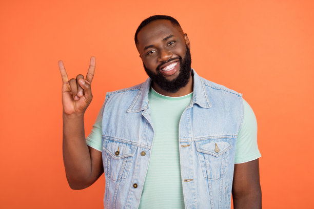 穿着牛仔裤和马甲的有角时尚男士在橙色背景下做出摇滚手势的照片图片下载