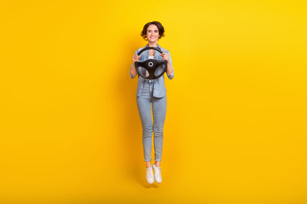 年轻女性在黄色背景下充满活力地跳起，手握方向盘，享受假期的全身照片图片下载