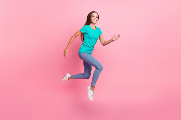 穿着青色T恤和牛仔裤的活力四射的女士在粉色背景上跳跃向前奔跑的照片图片下载