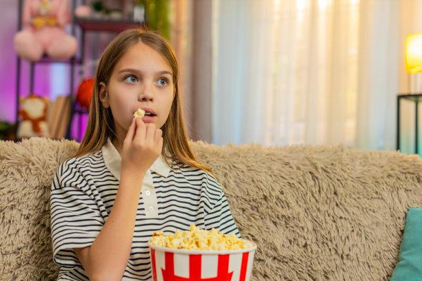 小女孩开心地吃着爆米花，一边看电视上的卡通，一边在沙发上快乐地笑着。图片下载