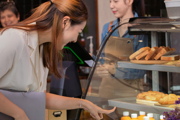 亚洲年轻女性小企业主在咖啡店为顾客使用POS收银机下单，咖啡师和服务员在舒适的工作环境中站在柜台后忙碌。图片下载