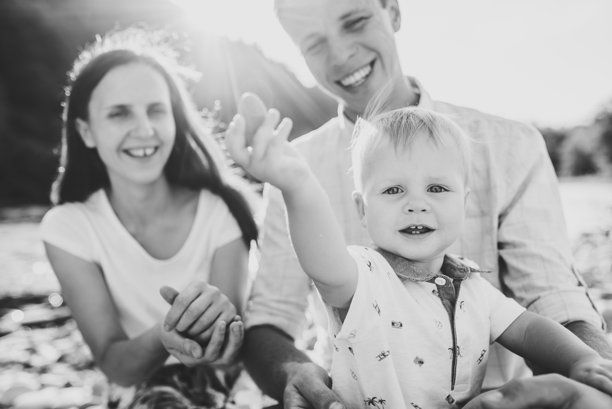 快乐的家庭夏日假期。母亲、父亲和婴儿儿子坐在湖边的沙滩上。父亲、母亲、儿童日。一起度过时光。妈妈和爸爸在日落时与孩子玩耍。黑白照片。图片下载