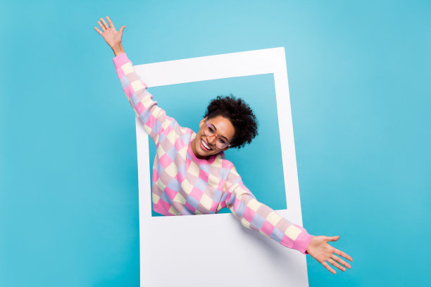 一个迷人快乐的女孩从照片框中走出来，向大家打招呼，享受乐趣，背景是明亮的蓝色。图片下载