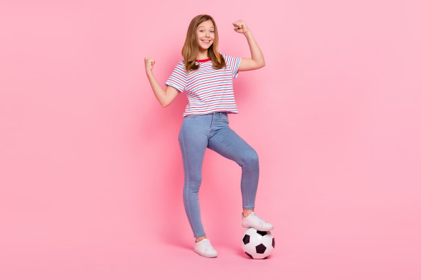 可爱的小金发女孩穿着T恤、牛仔裤和鞋子在粉色背景上踢足球的全身照片图片下载