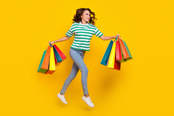 可爱的棕发千禧女性穿着T恤、牛仔裤和鞋子，手提购物袋在黄色背景上奔跑的全身照片图片下载