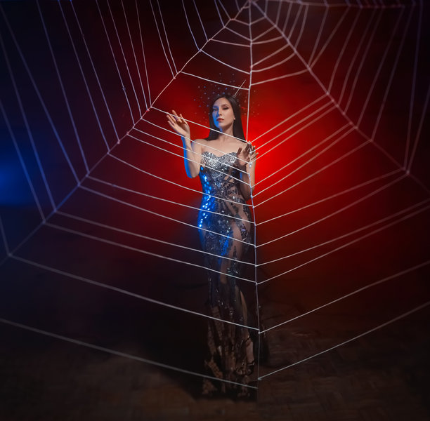 穿着闪亮银色裙子的蜘蛛女王幻想照片，双手触摸着巨大的蛛网。狂欢节服装黑寡妇王冠。女孩公主时尚模特在红色灯光背景下摆姿势。图片下载