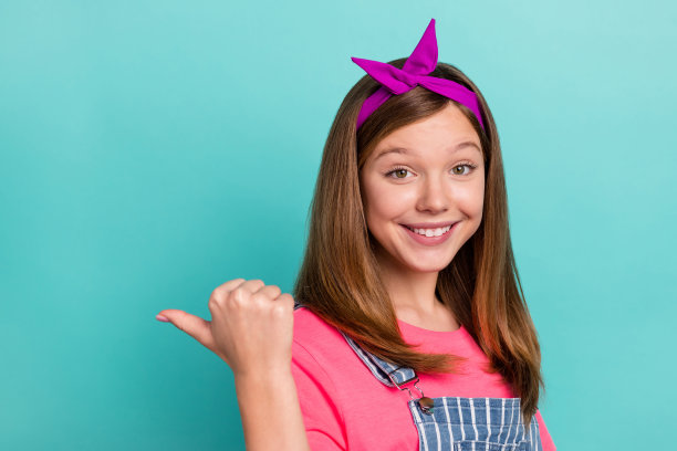 可爱的棕色头发小女孩的个人头像，戴着发带，穿着粉色T恤和背带裤，背景为青色。图片下载