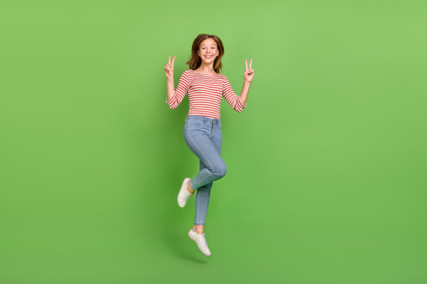 穿着条纹衬衫、牛仔裤和运动鞋的快乐友好的女士在绿色背景前跳跃并做出V字手势的照片图片下载