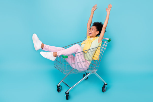 年轻女士在购物中心骑购物车，享受乐趣的全身侧面照片，背景为蓝色。图片下载