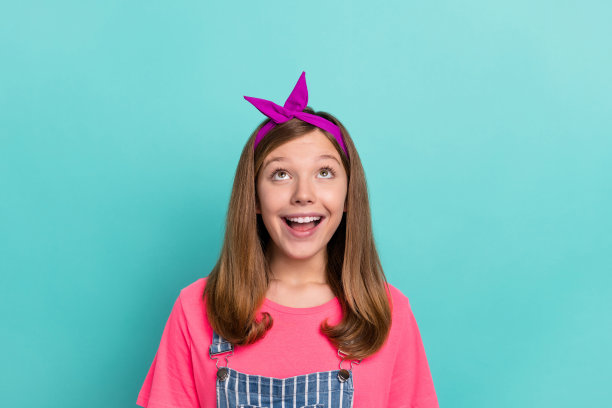 一位印象深刻的棕发少女，佩戴发带，穿着粉色T恤和背带裤，孤立在青绿色背景前仰望。图片下载