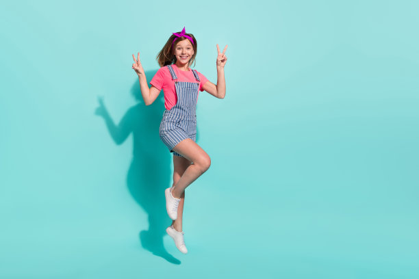 全身照片，女孩跳跃做V字手势，穿着条纹T恤和短裤，背景为青色。图片下载