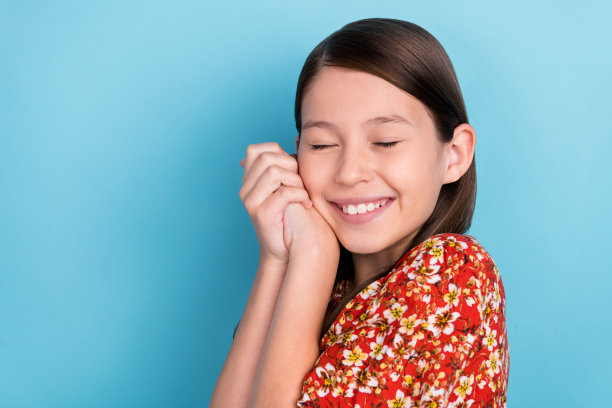 侧面肖像照片，年轻快乐的女孩享受梦想，双手轻触脸颊，背景为蓝色。图片下载