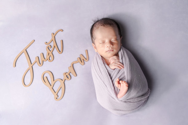 一张专业的两周大日本婴儿照片，婴儿被柔软的浅紫色毛毯包裹，安静地睡着。旁边轻轻放着一个写有“刚出生”的木牌。图片下载